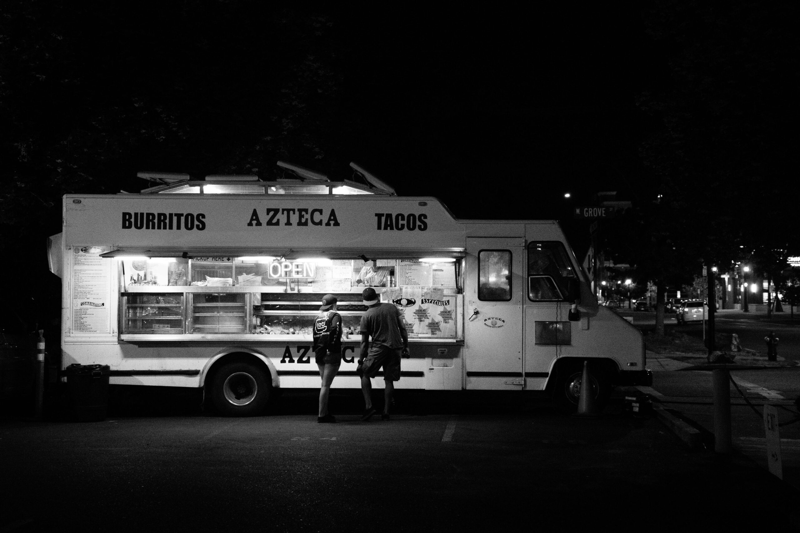 Home Black and white photo of people buying from a taco food truck at night.