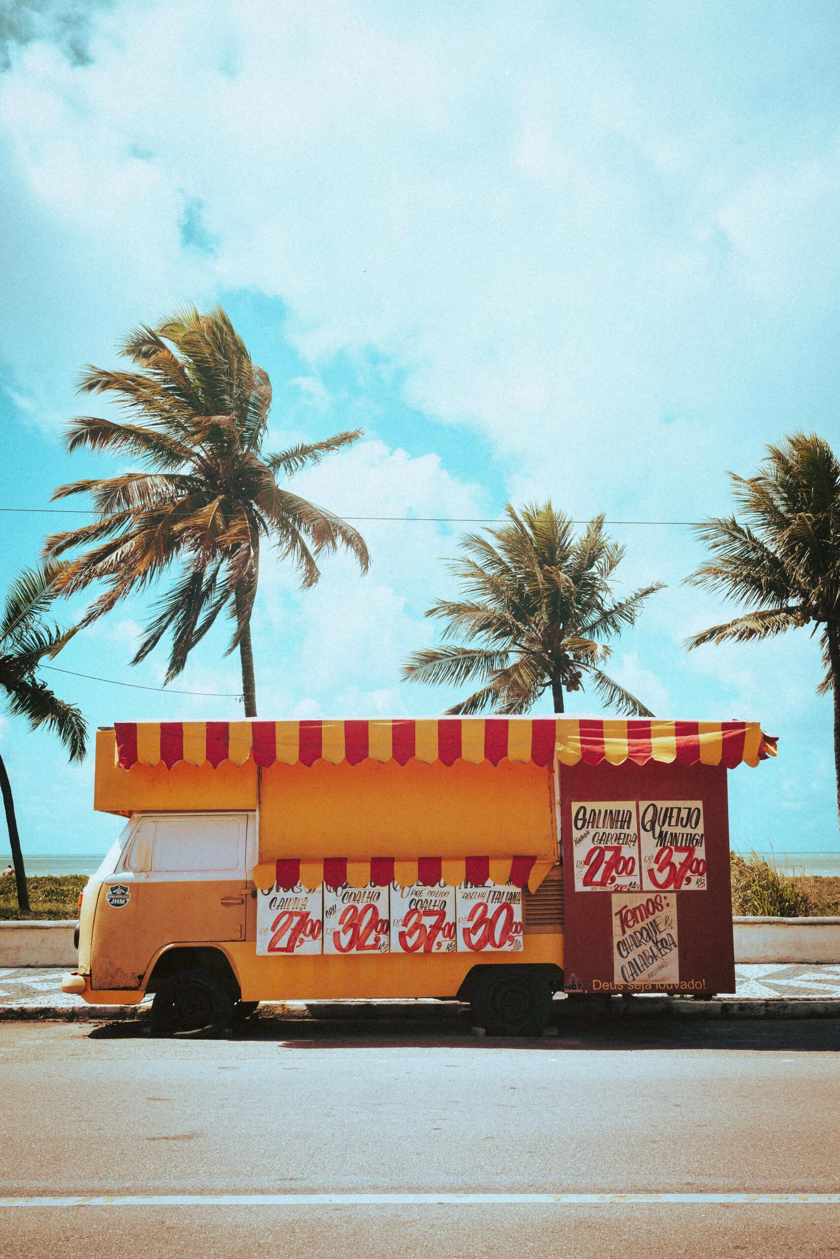 Home A colorful food truck parked under swaying palm trees on a sunny urban street.