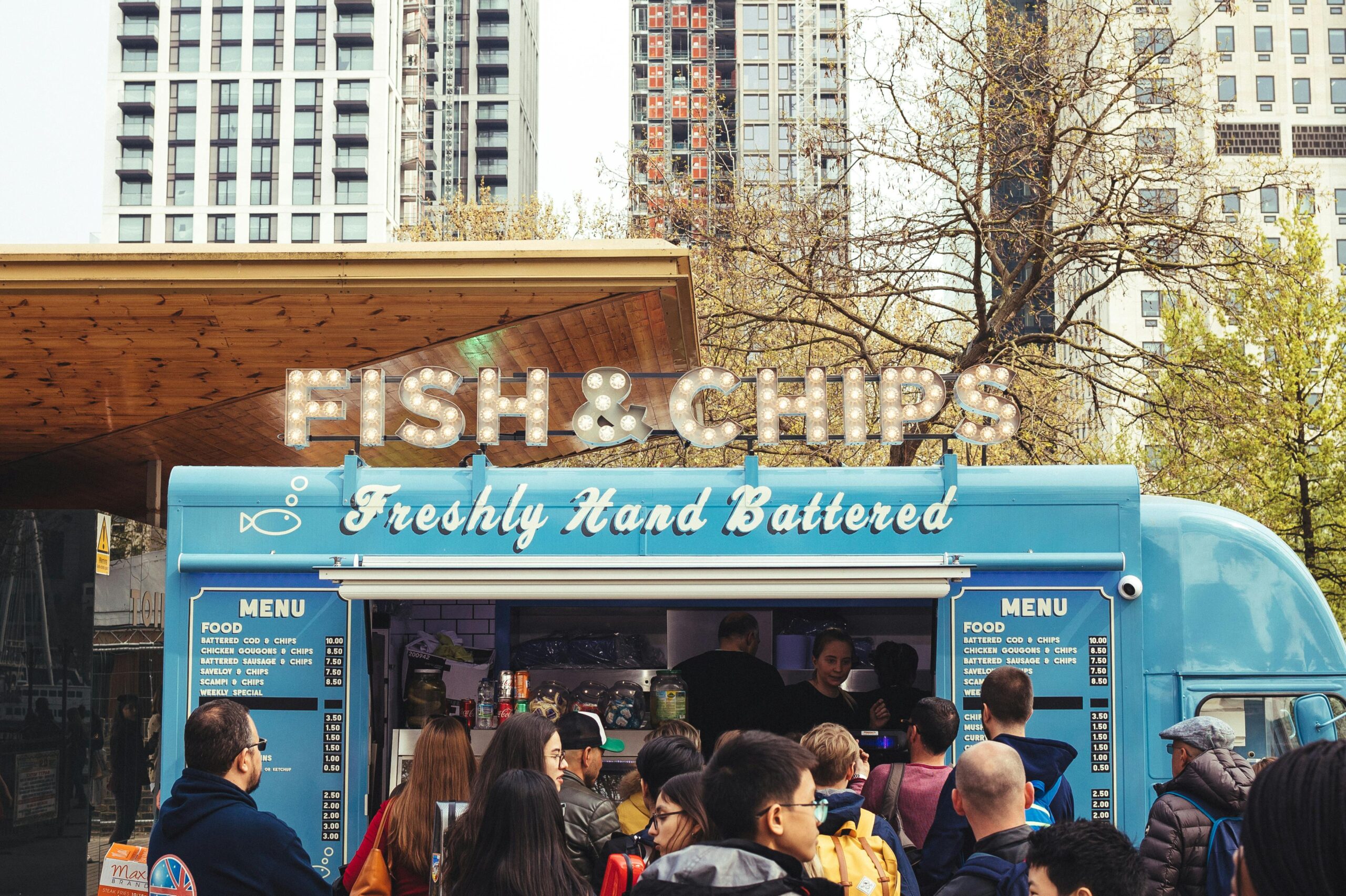 Home Crowd enjoying fish and chips at a lively food truck with urban buildings in the background.